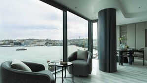 Una sala de estar moderna con ventanas amplias, vista a un paisaje urbano y al agua, y un comedor con mesa y sillas.