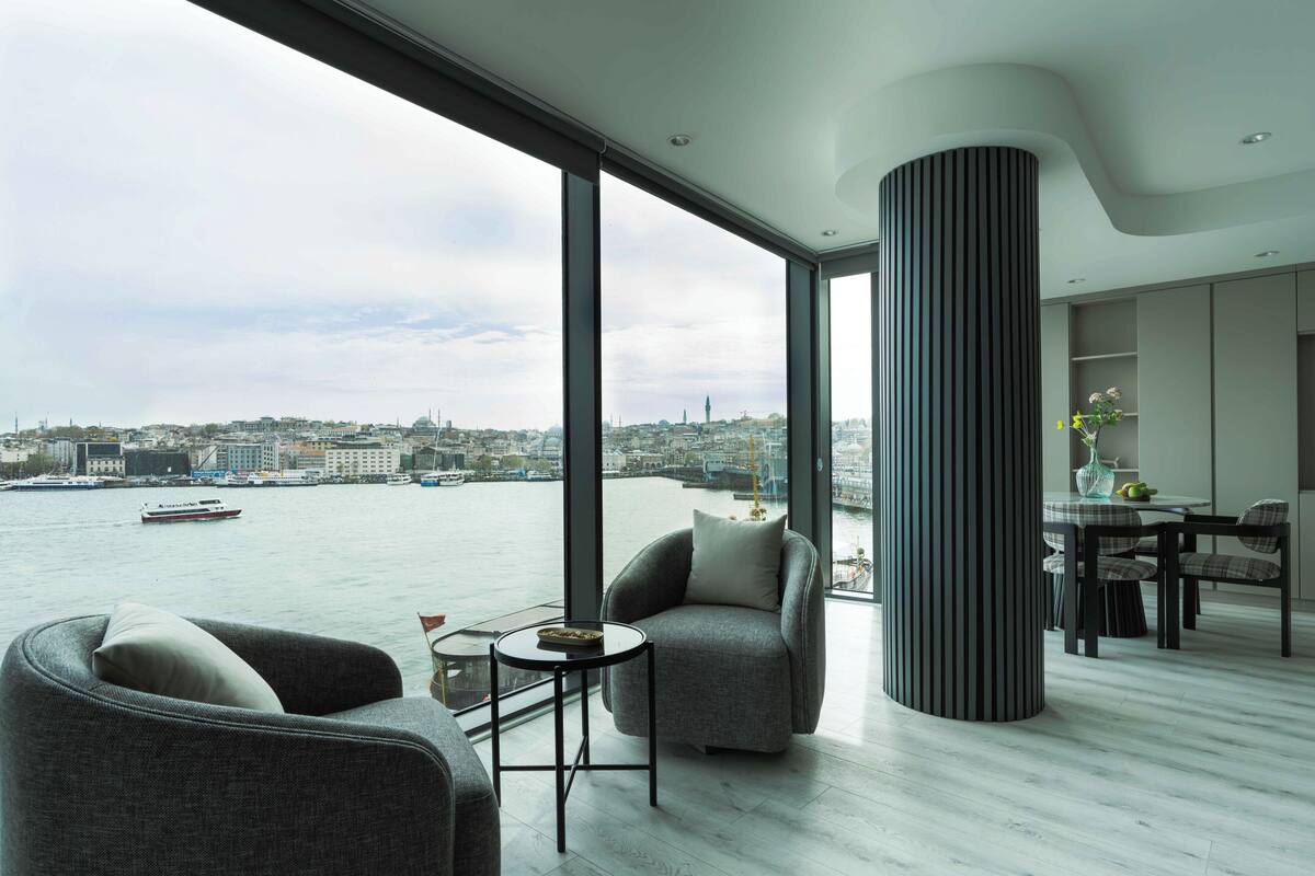 Una sala de estar moderna con ventanas amplias, vista a un paisaje urbano y al agua, y un comedor con mesa y sillas.