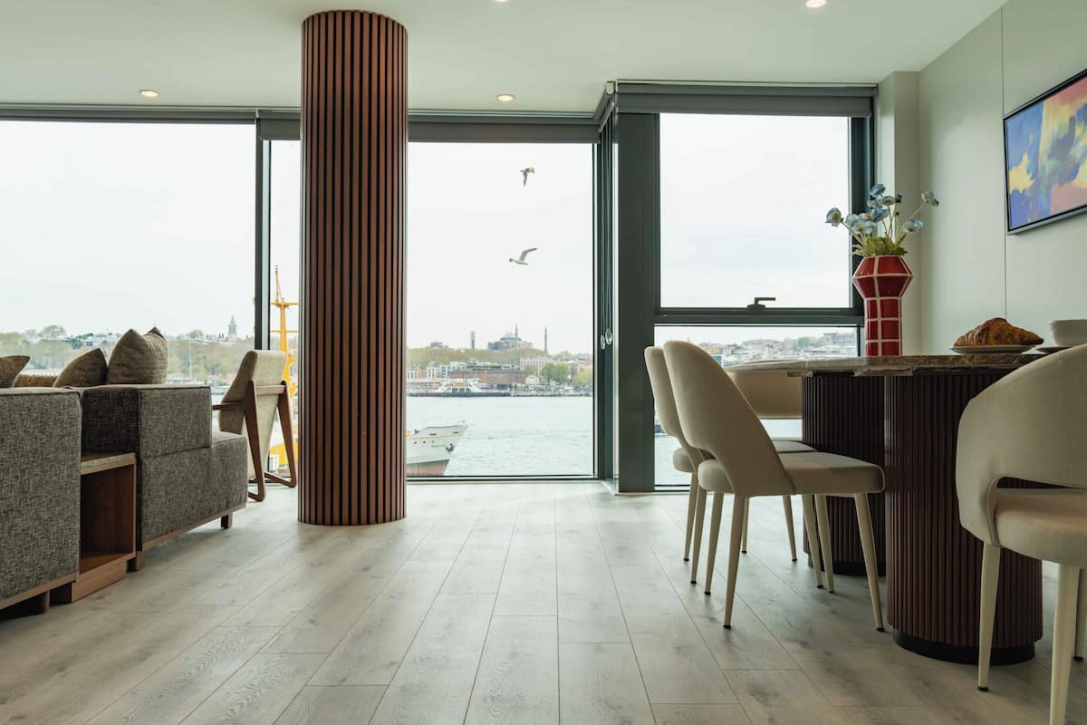 Un salón moderno con un ventanal grande con vistas a un paisaje urbano, un pilar de madera y un comedor con sillas y una mesa.
