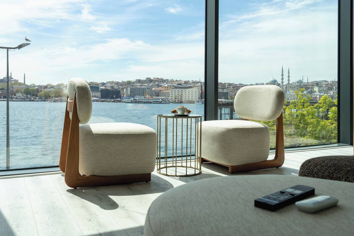 Habitación moderna con dos sillones tapizados, una mesita con taza y platillo, y un control remoto sobre una superficie de madera. Un ventanal grande ofrece vistas a un paisaje urbano y una masa de agua.