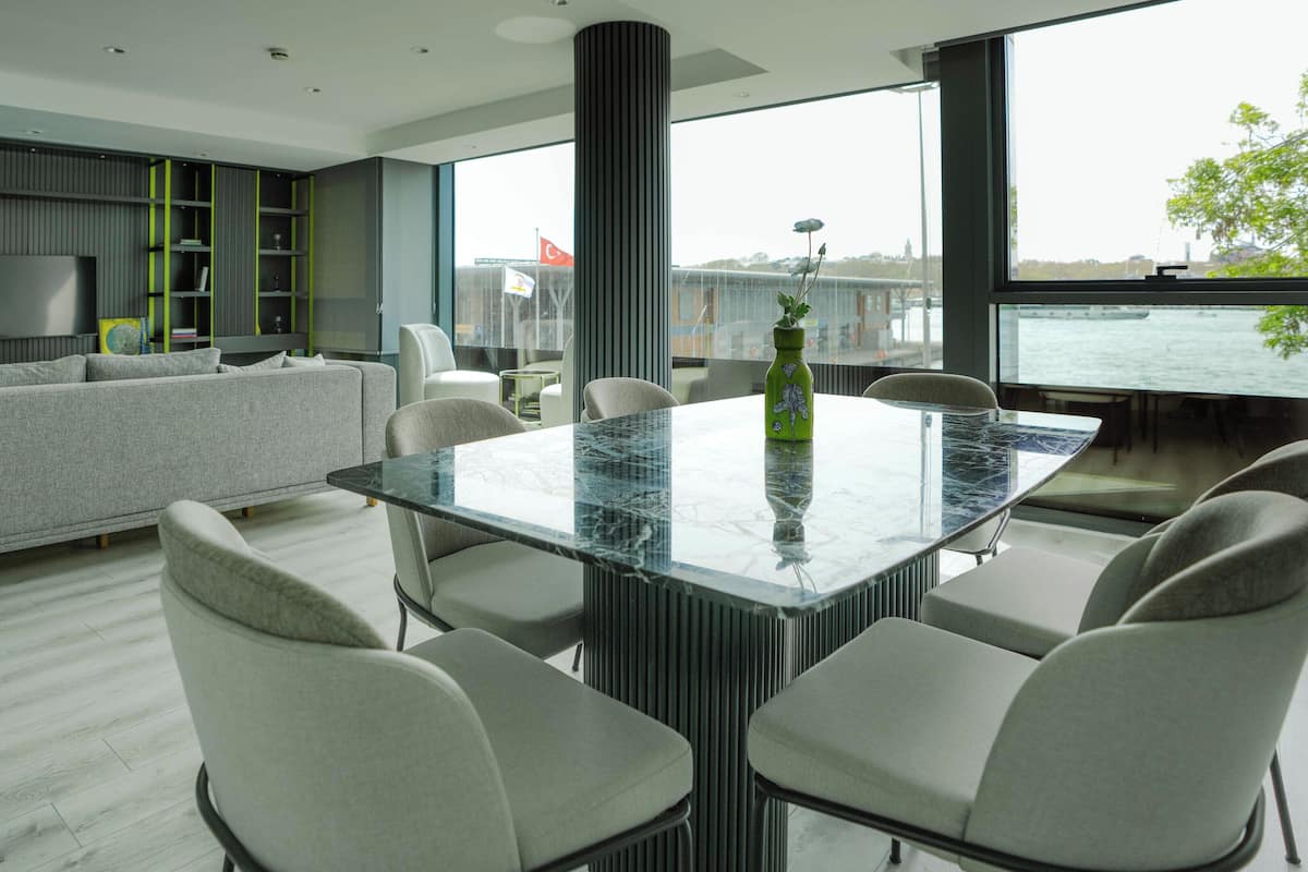 Una sala de estar moderna con un sofá gris, una mesa de comedor con superficie de vidrio y vistas a una masa de agua a través de amplios ventanales.