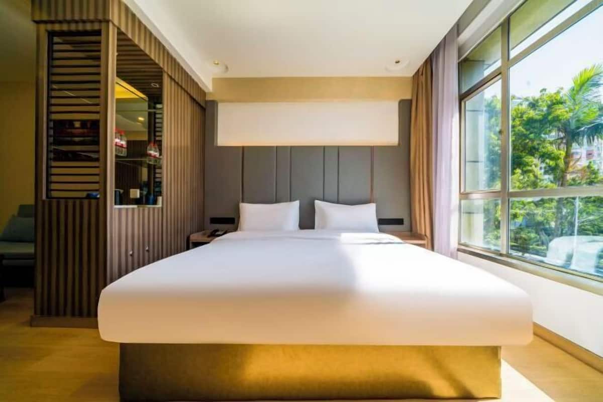 A modern hotel room with a large bed, a nightstand, a wardrobe, and a large window offering a view of greenery outside.