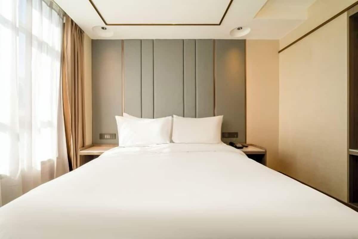 A hotel room with a large bed, a headboard with vertical lines, a bedside table with a lamp, and a window with curtains.