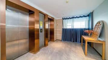 An elevator area with metallic doors, a wooden bench, and a window with blue curtains.