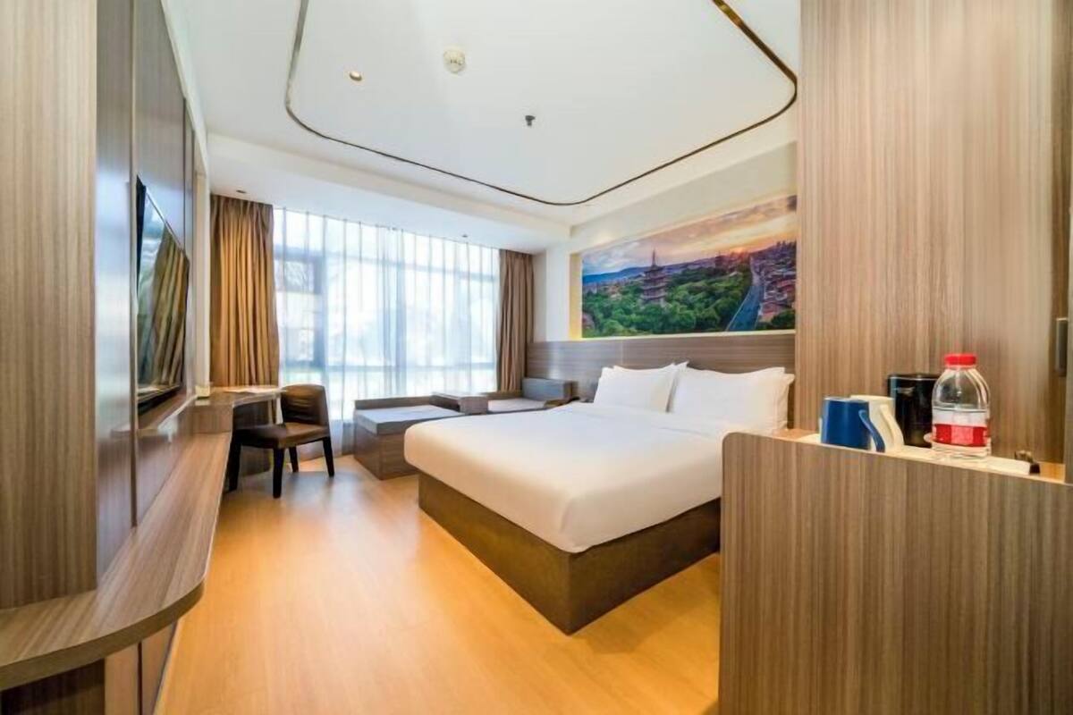 A modern hotel room with a large bed, a desk, and a view of a cityscape through the window.