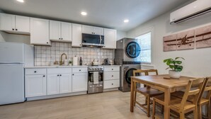 A modern kitchen with white cabinets, a microwave, oven, and washing machine. There is a dining table with chairs and a potted plant.