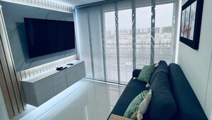 A modern living room with a flat-screen TV mounted on a wall, a sofa with cushions, a wooden coffee table, and a large window with a city view.