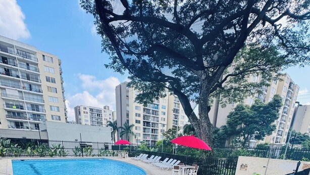 A swimming pool with lounge chairs and umbrellas, surrounded by greenery and tall buildings in the background.