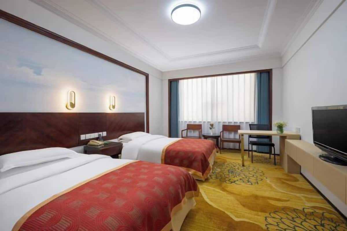 A hotel room with two beds, a desk with a chair, a television, and a large window with curtains.