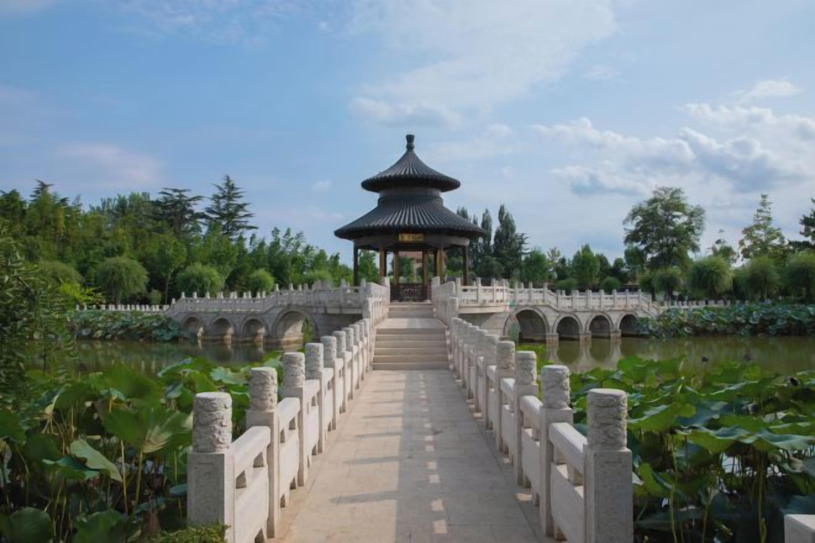 A traditional pavilion with a multi-tiered roof, a stone bridge leading to it, and a calm water body with lotus plants.