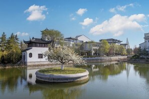 A traditional building with a pond in front, surrounded by trees and a clear sky. - Qingdaojinxiushui Hotel (Pingdu)