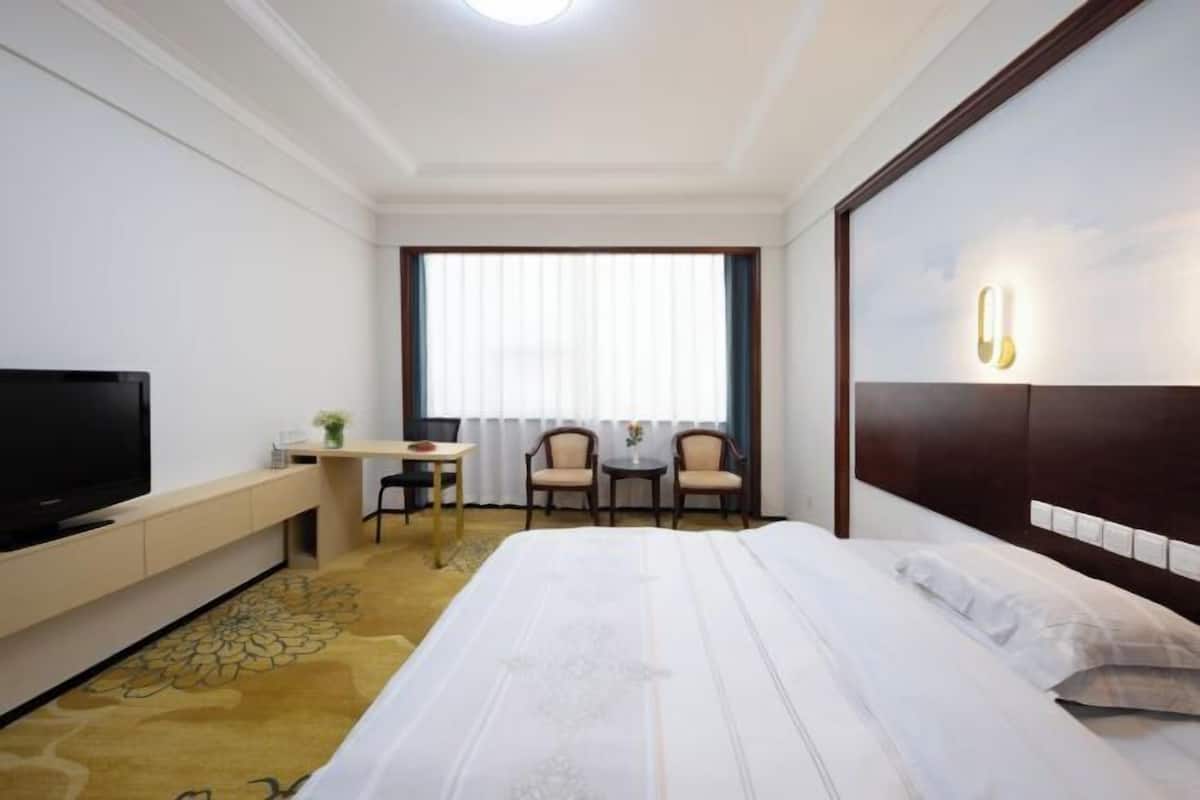A hotel room with a bed, TV, desk, chairs, and a window with curtains.