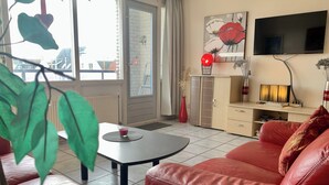 Un salon avec un canapé en cuir rouge, une table basse, un téléviseur fixé au mur et une fenêtre donnant sur des bâtiments.