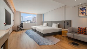 A modern hotel room with a large bed, a desk, a chair, a sofa, and a large window offering a city view.