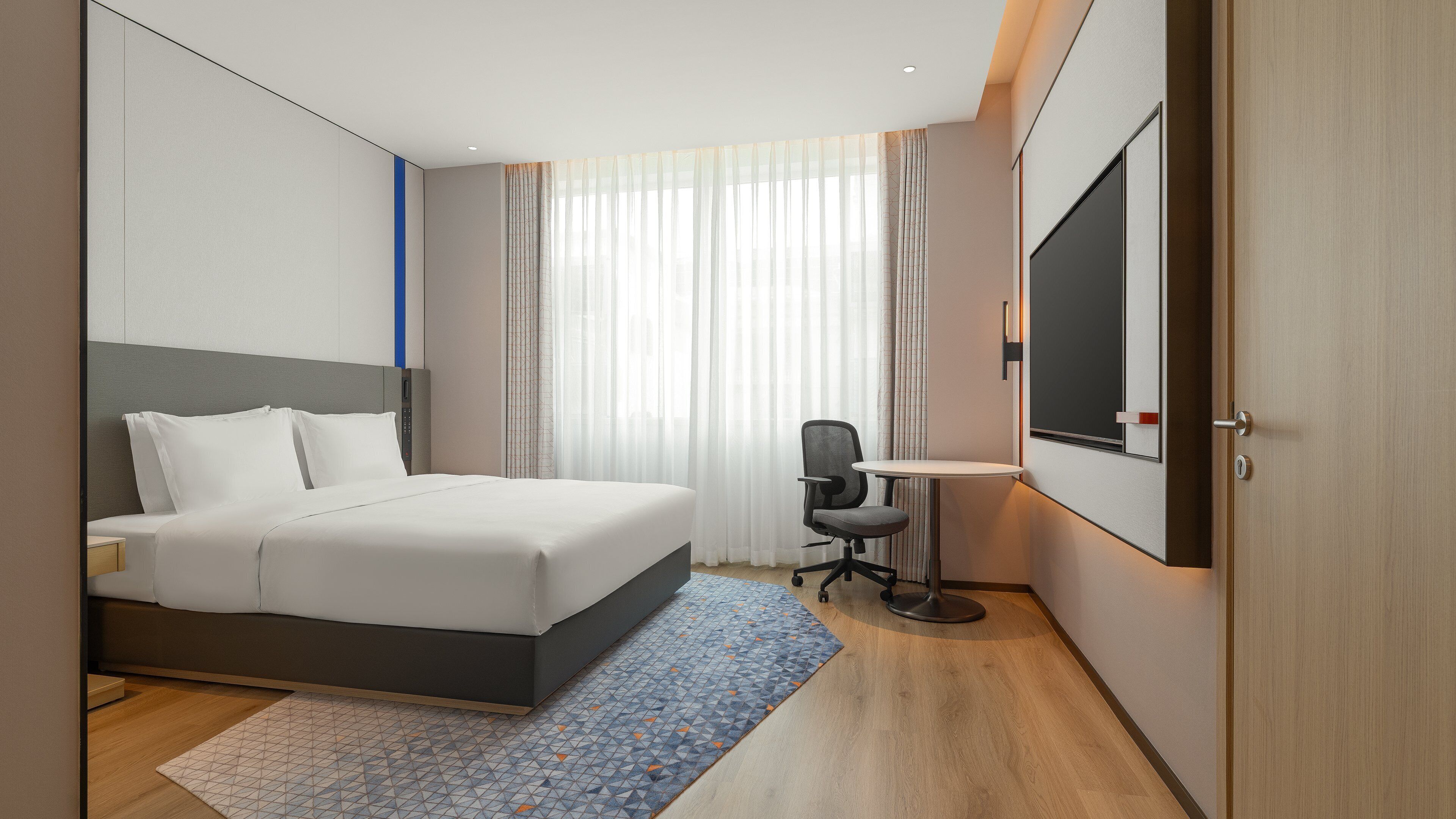 A hotel room with a bed, a desk, a chair, a TV, and a door.