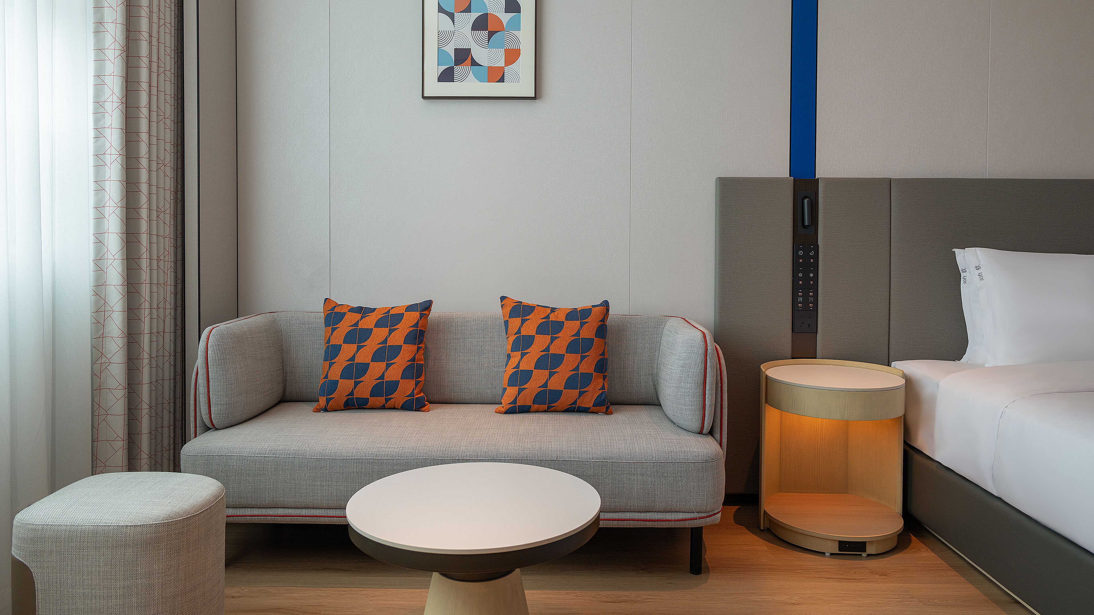 A modern hotel room with a sofa, two patterned pillows, a small round table, a lamp, and a bed.