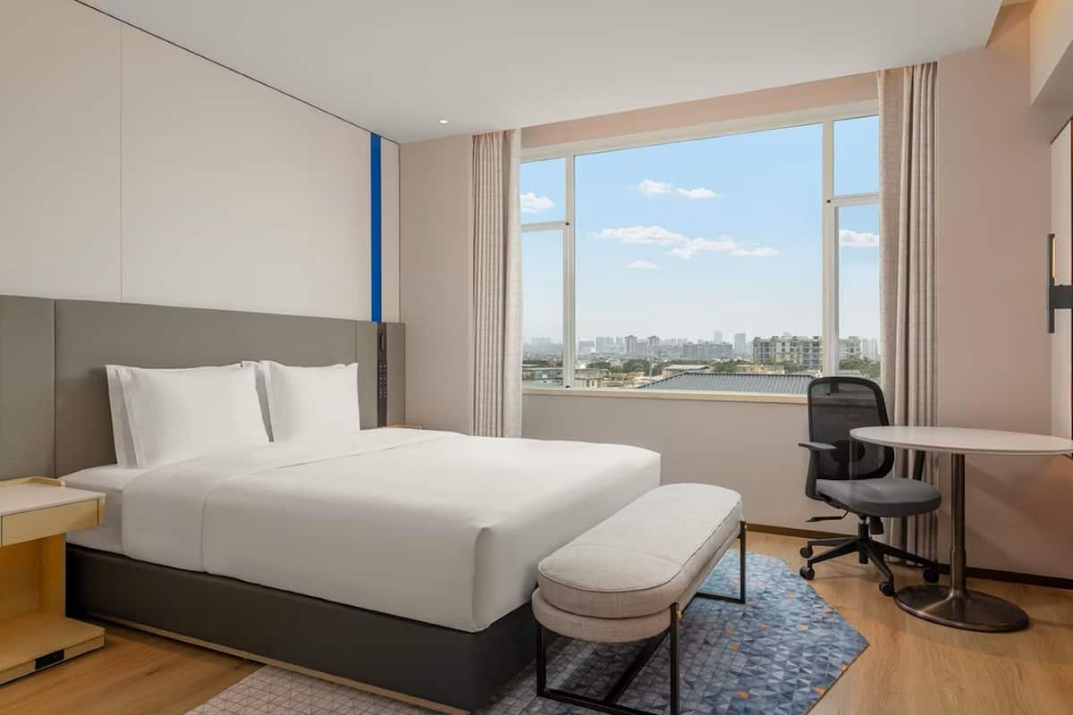 A modern hotel room with a large bed, a desk, a chair, and a view of the cityscape.