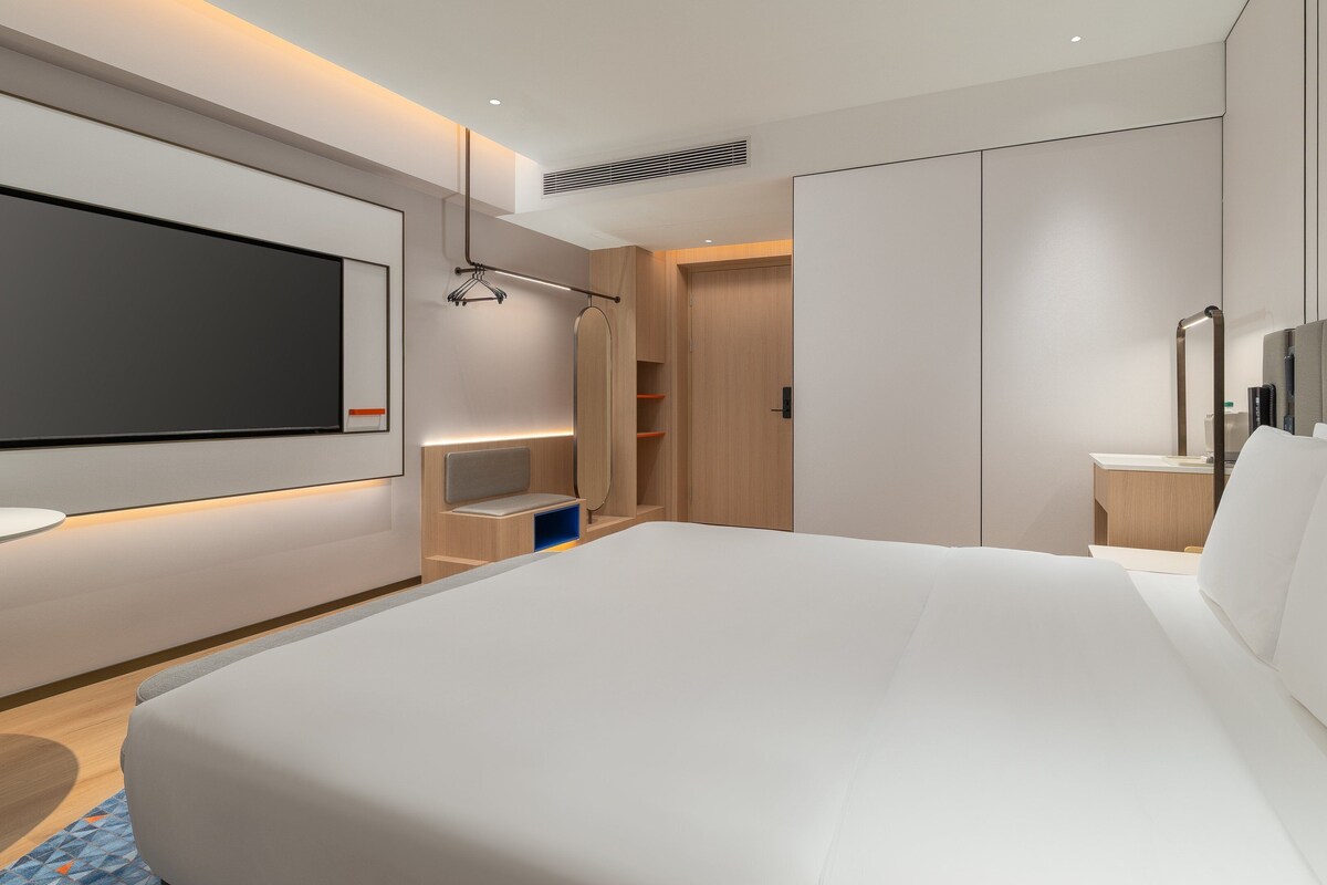 A modern hotel room with a large bed, a TV mounted on the wall, a bedside table, and a wardrobe.