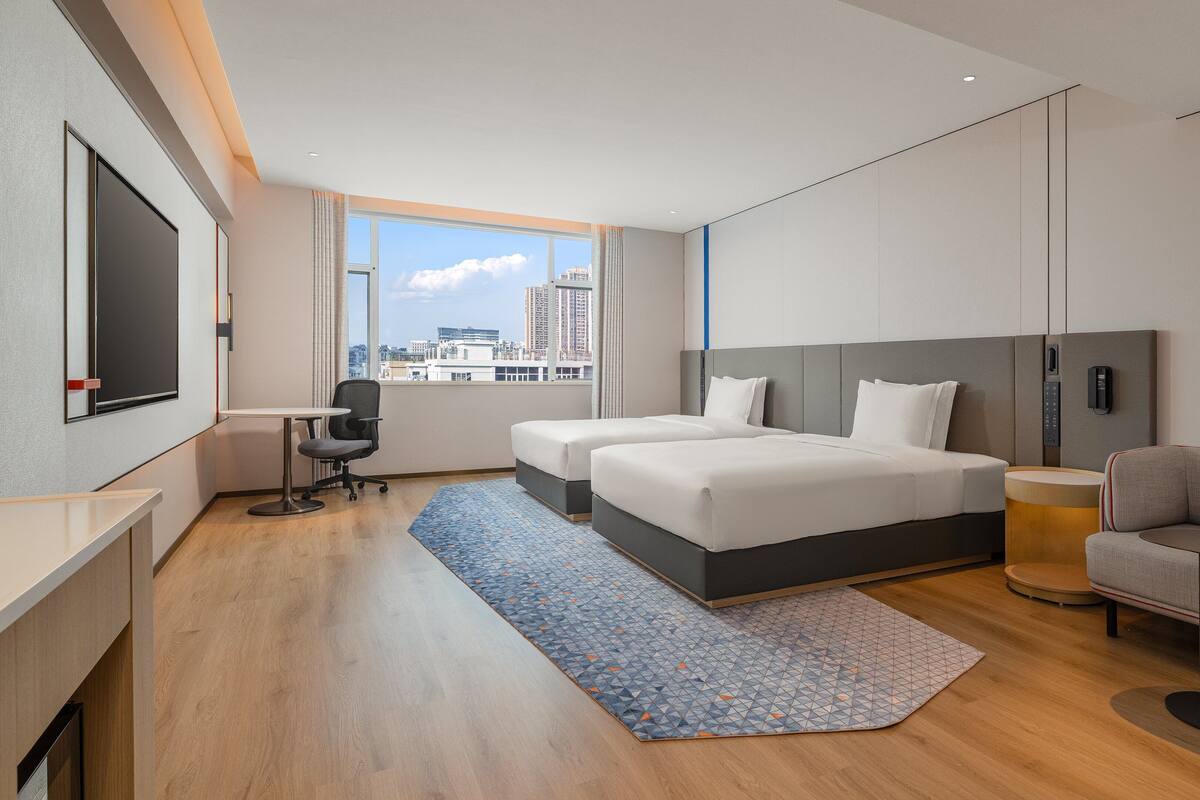 A hotel room with a large bed, a desk, a chair, a sofa, and a city view.