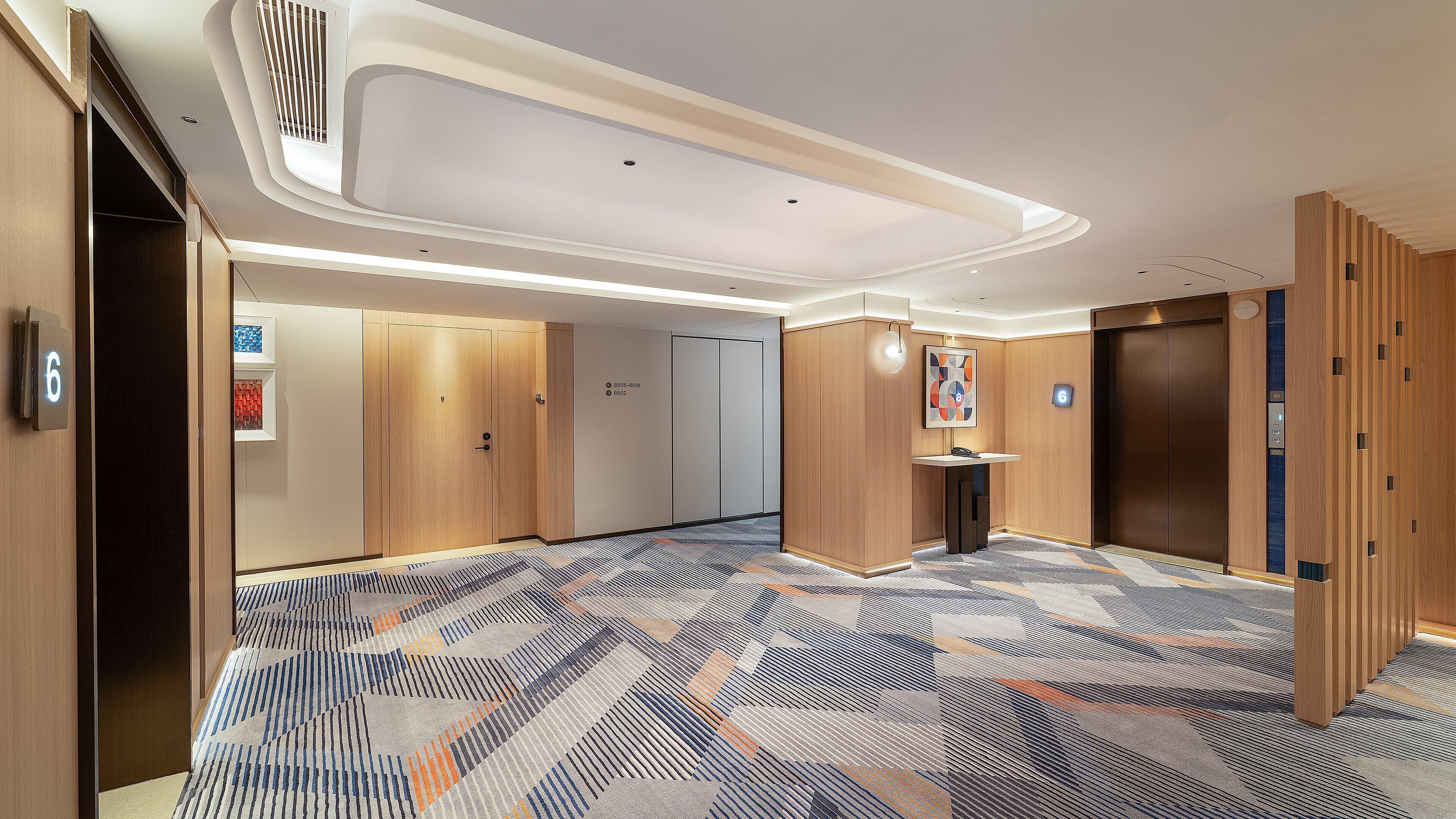 Un couloir d’hôtel avec un tapis à motifs, des portes en bois, une petite table avec un téléphone et un ascenseur.