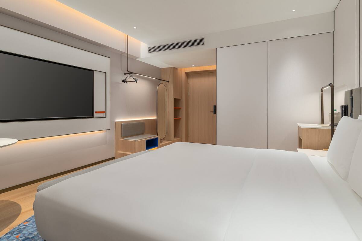 A modern hotel room with a large bed, a TV mounted on the wall, a bedside table, and a wardrobe.