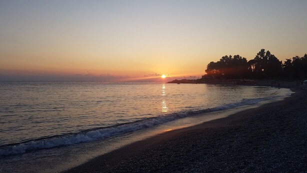 A beach at sunset with calm waves, a clear sky, and trees in the background. - Cozy beach house 150 mt from the beach kitchen and shower also outside (Nocera Scalo)