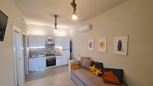 A modern kitchen with white cabinets and a tiled backsplash, a grey sofa with orange cushions, and wall art depicting a wave, sun, and surfboard. - Cozy beach house 150 mt from the beach kitchen and shower also outside (Nocera Scalo)
