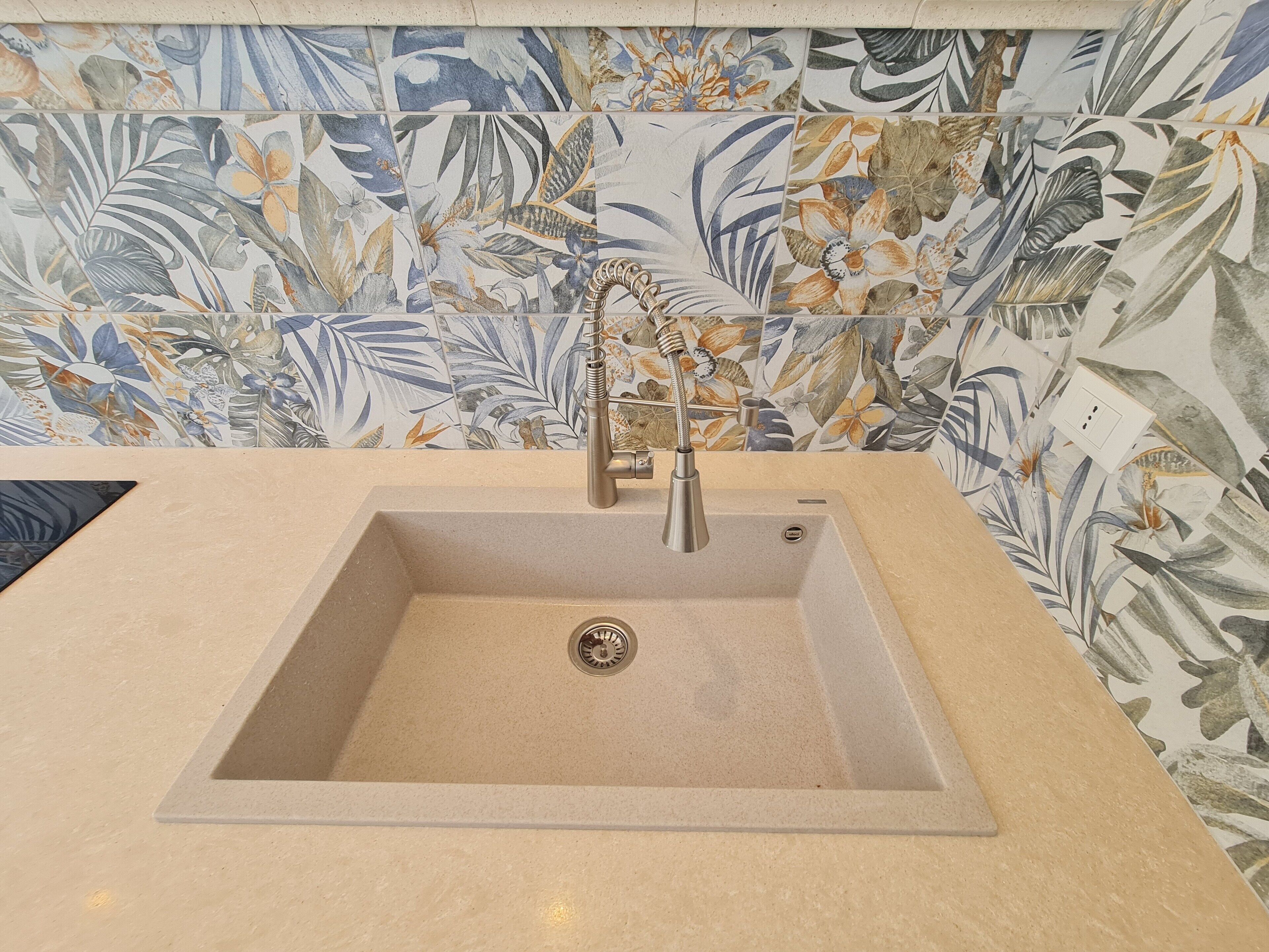 A bathroom with a beige sink, a modern faucet, and a decorative tile backsplash featuring tropical leaf patterns.
