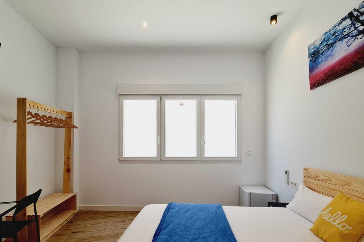 A bedroom with a bed, a bench, a desk, a chair, a window, and a painting on the wall.