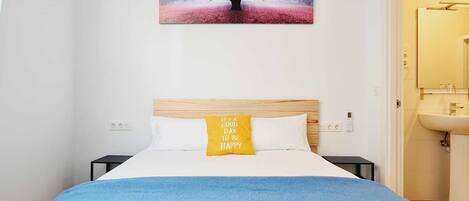 Un dormitorio con cama, manta azul, almohada amarilla, cabecera de madera y un cuadro de un árbol en la pared.