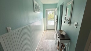 A hallway with light blue walls, a wooden floor, a white radiator, a door with a view to the outside, and a small table with a mirror and a shelf. - Island Time - 2 Minute Walk To The Beach & Heated Pool (Holmes Beach)