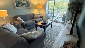 A living room with a gray sofa, a coffee table, a lamp, and a painting of sea turtles. - Island Time - 2 Minute Walk To The Beach & Heated Pool (Holmes Beach)