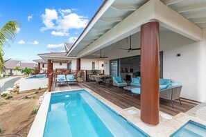 Un área de piscina moderna al aire libre con un deck de madera, sillones de descanso y un patio cubierto.