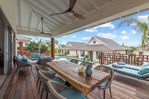Un espacio exterior cubierto con piso de madera, mobiliario de mimbre, piscina y vista a casas vecinas.