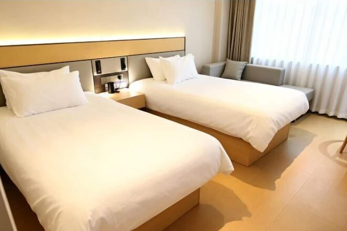 A hotel room with two beds, a bedside table, a lamp, and a window with curtains.