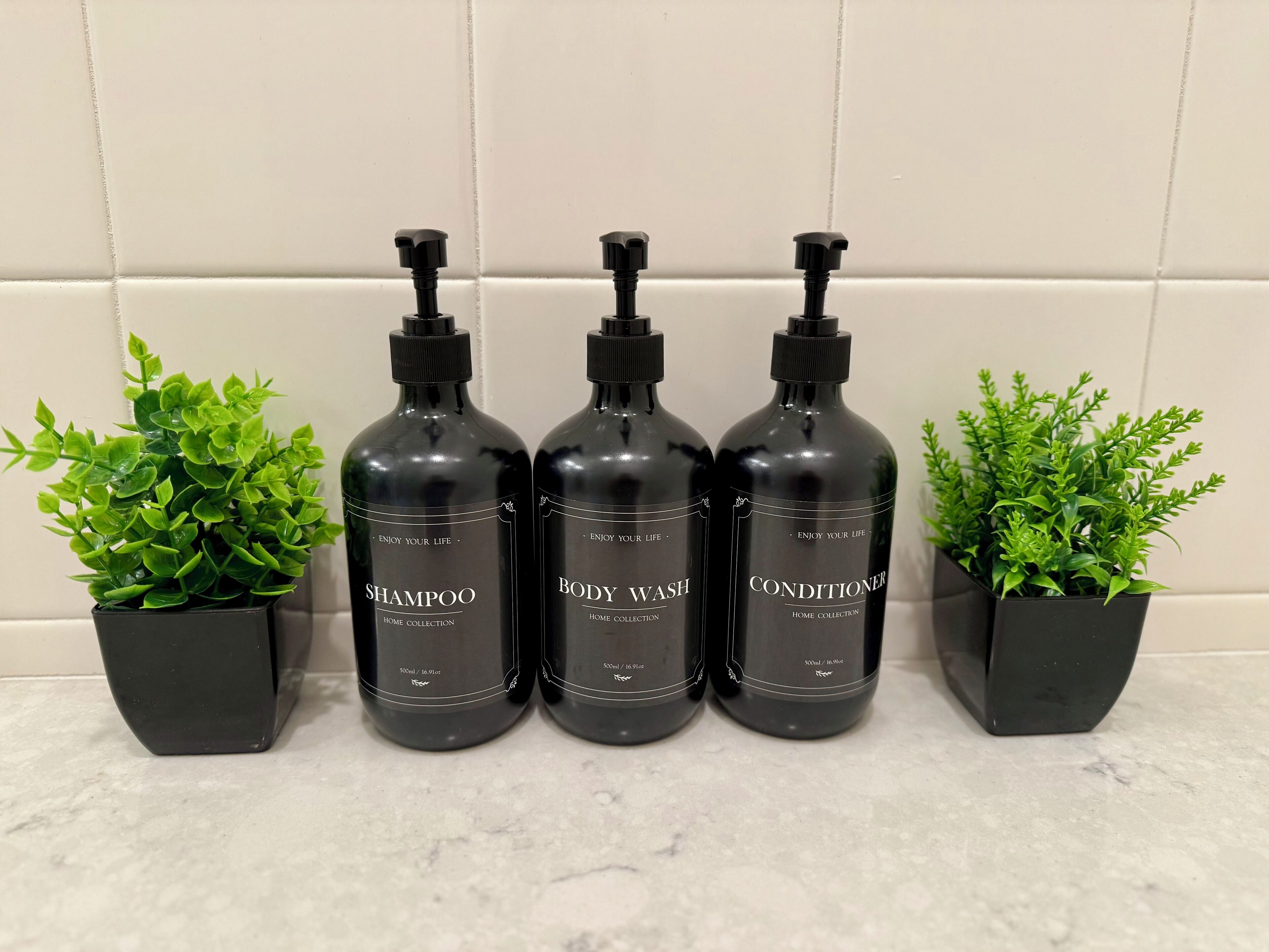 Trois flacons noirs contenant du shampoing, du gel douche et de l’après-shampoing, posés sur un comptoir carrelé.