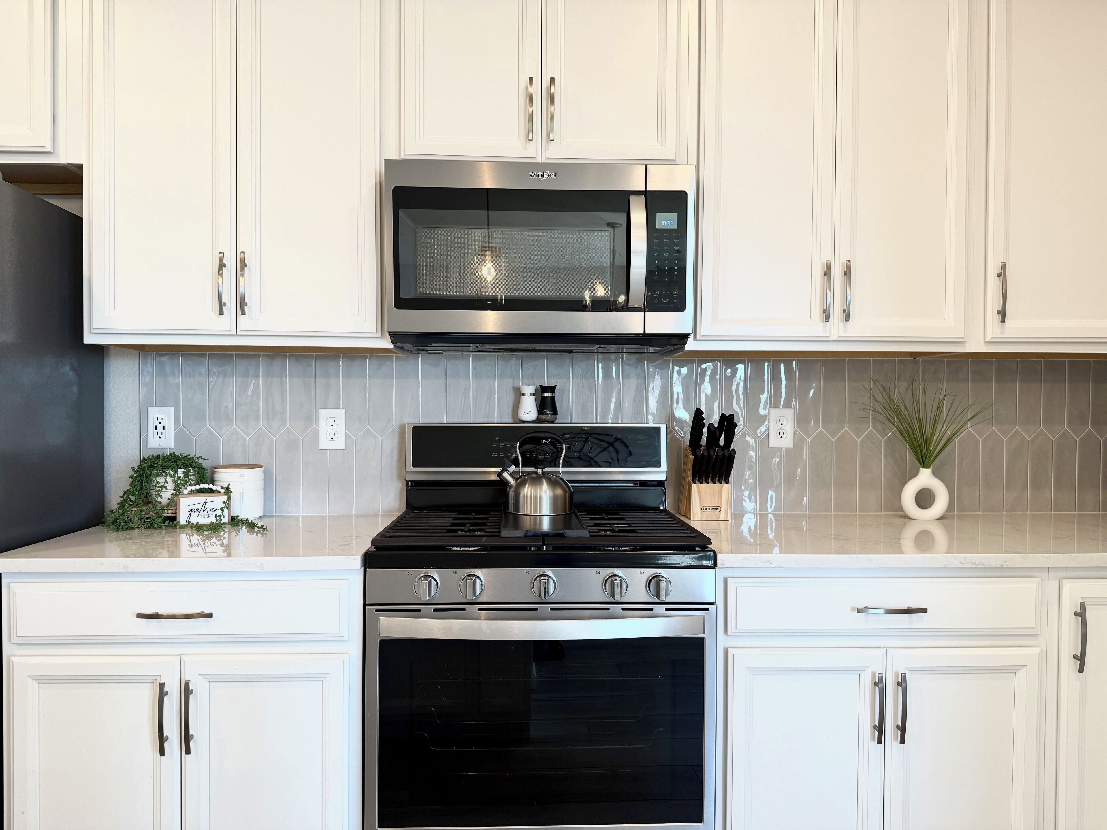 Cozinha moderna com armários brancos, fogão de aço inoxidável, micro-ondas e painel de azulejos.