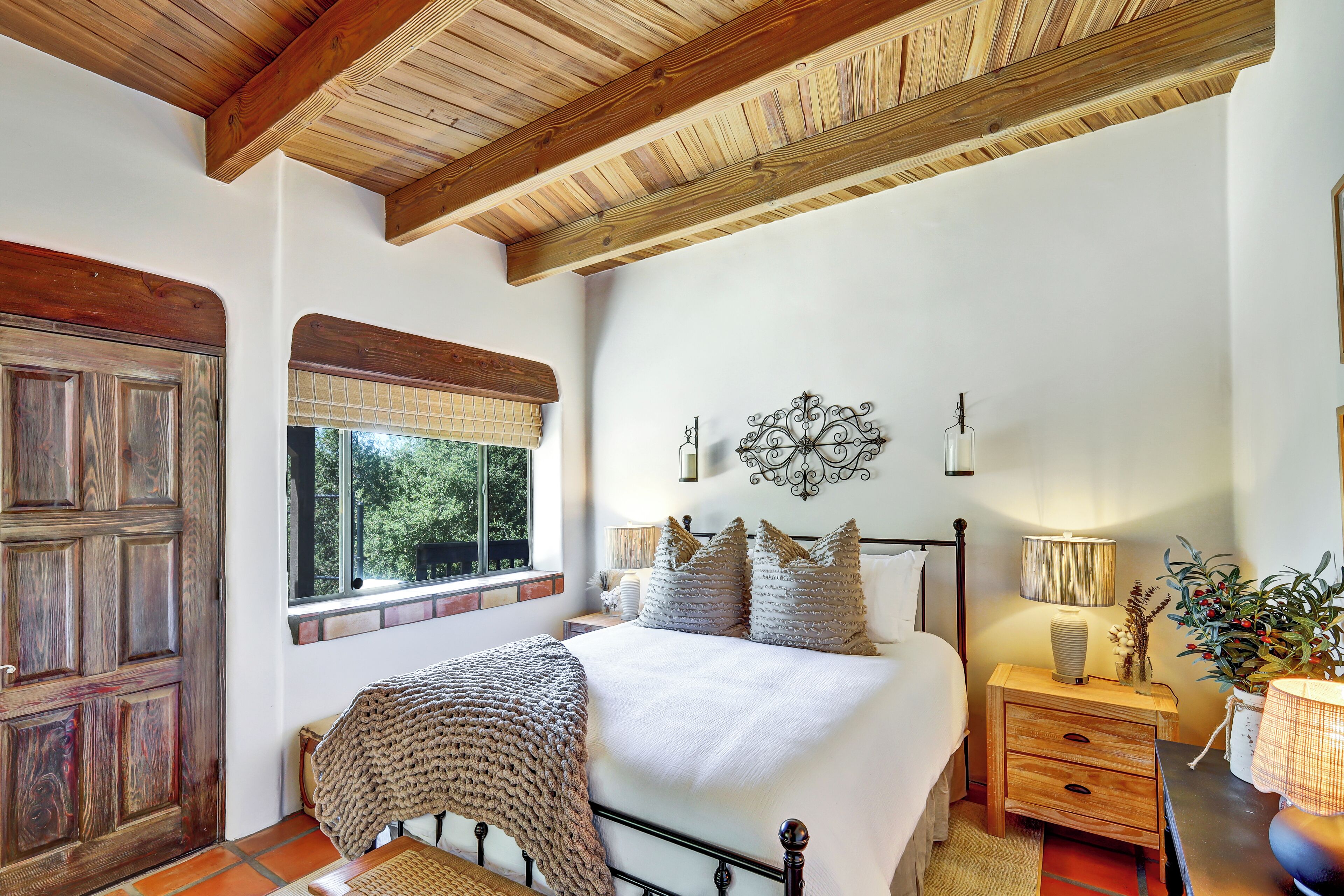 A bedroom with a wooden ceiling, a large bed with white bedding, a nightstand with a lamp, and a window with a view of greenery.