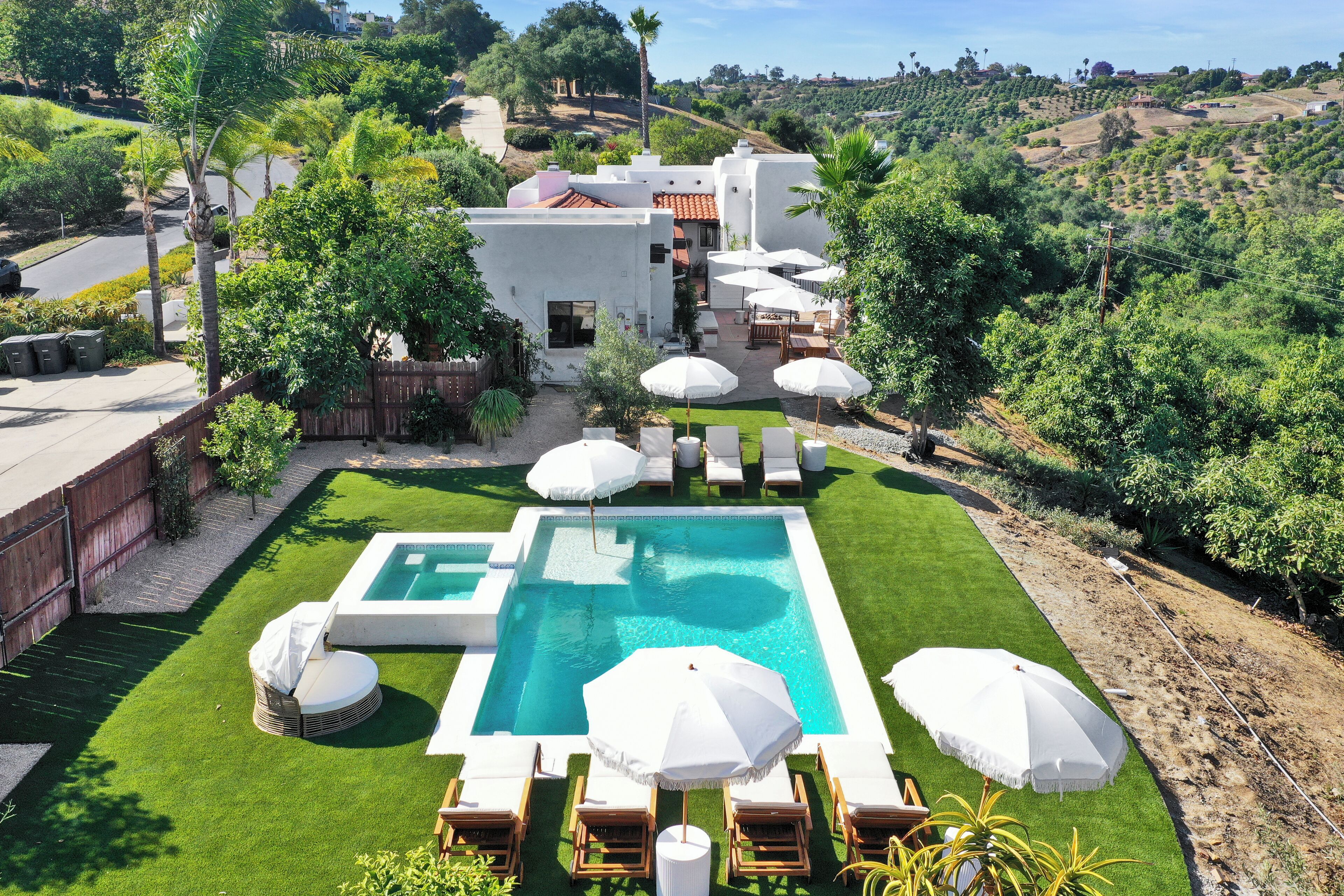 A pool area with a rectangular pool, surrounded by white lounge chairs and umbrellas, set against a backdrop of lush greenery and a hilly landscape.
