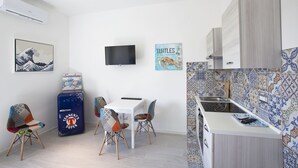 Ett kompakt kök med kakel på väggen bakom arbetsbänken, en platt-TV som är monterad på väggen och en liten matplats med ett bord och stolar.