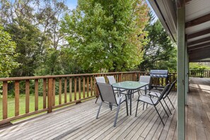 A wooden deck with a glass-top table and chairs, a grill, and a wooden railing.