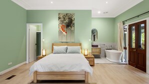 A bedroom with a bed, bedside tables, a nightstand, a large painting, a bathroom with a bathtub, and a door leading outside. - Birdstow – Spacious, peaceful Stay, Sleeps 10 (Wentworth Falls)