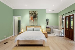 A bedroom with a bed, bedside tables, a nightstand, a large painting, a bathroom with a bathtub, and a door leading outside. - Birdstow – Spacious, peaceful Stay, Sleeps 10 (Wentworth Falls)