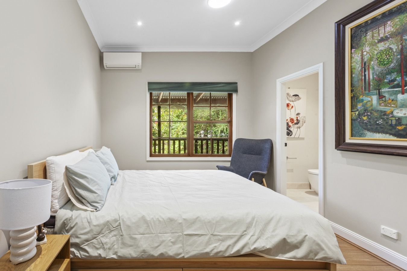 Een slaapkamer met een bed, een stoel, een raam met uitzicht op groen en een schilderij aan de muur.