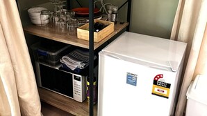 A compact kitchen with a white refrigerator, a wooden shelf unit with dishes and glasses, and a small storage unit. - Sakura – Calm, Cozy, & Close to Nature (Wentworth Falls)