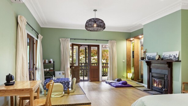 A cozy room with a wooden floor, a dining table with chairs, a sofa, a fireplace, and a large window with curtains. - Sakura – Calm, Cozy, & Close to Nature (Wentworth Falls)
