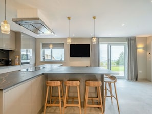 A modern kitchen with a central island, bar stools, pendant lights, a large window, and a flat-screen TV.