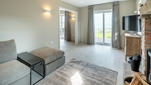En moderne stue med sofa, fjernsyn, pejs og udsigt gennem store vinduer.