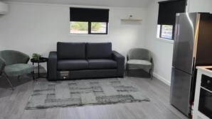 A modern living room with a grey sofa, two armchairs, a small side table with a plant, and a grey rug. There are two windows with black blinds, a built-in oven, and a stainless steel refrigerator. - Microtel Lodge (Hamilton)