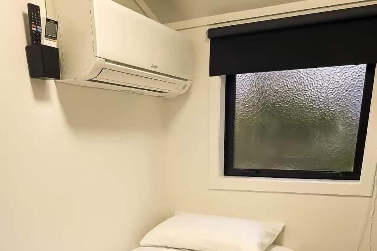 A single bed with white bedding and a rolled towel, an air conditioning unit above, a window with a black blind, and a wooden floor.